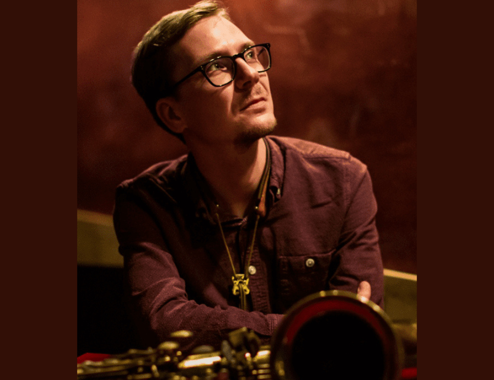 A musician wearing glasses sits pensivly with his saxaphone infront of them