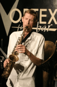 A man plays a saxophone in front of a "VORTEX" sign.
