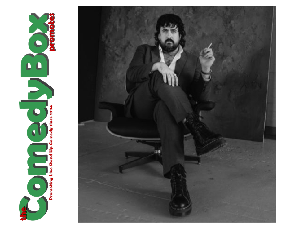 A person with dark curly hair sits in a chair holding a cigarette, wearing a suit and boots.
