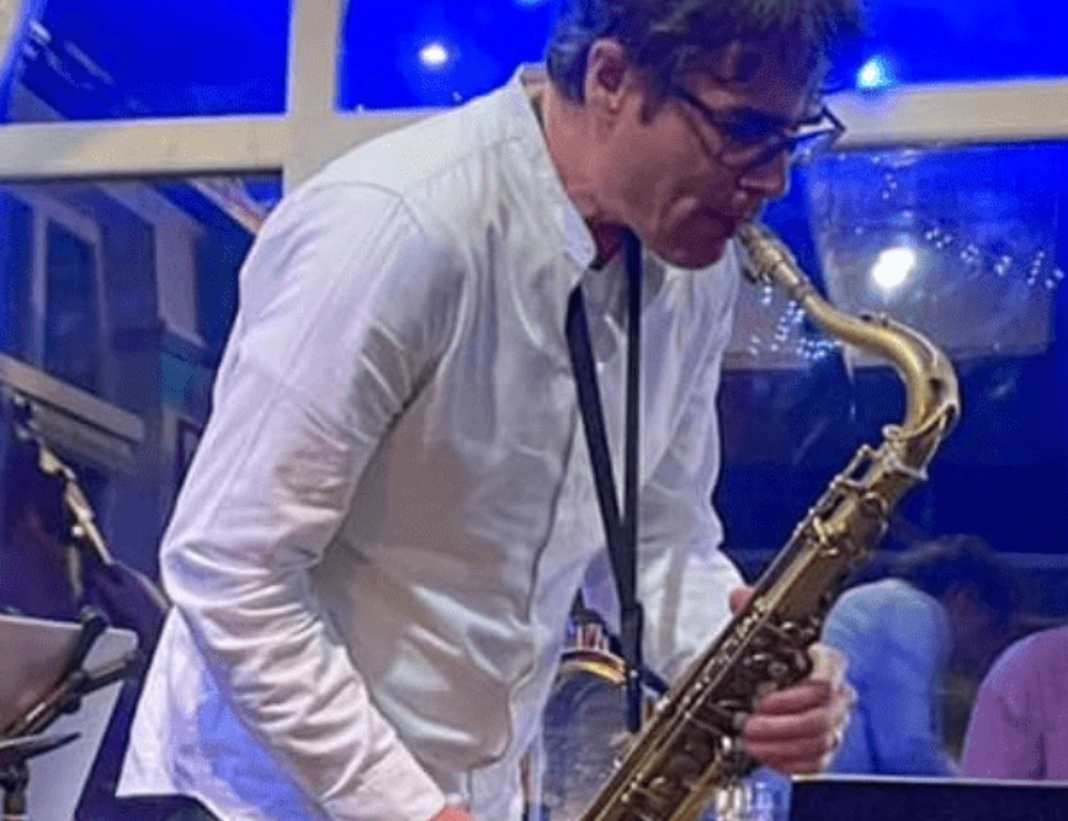 A man wearing glasses and a white shirt plays a saxophone. He is in focus while the background is blurred with blue lights.